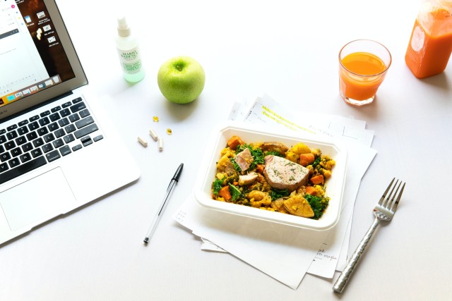 Een bureau met daarop een lunchsalade en bestek en daarnaast staat een opgeklapte laptop aan